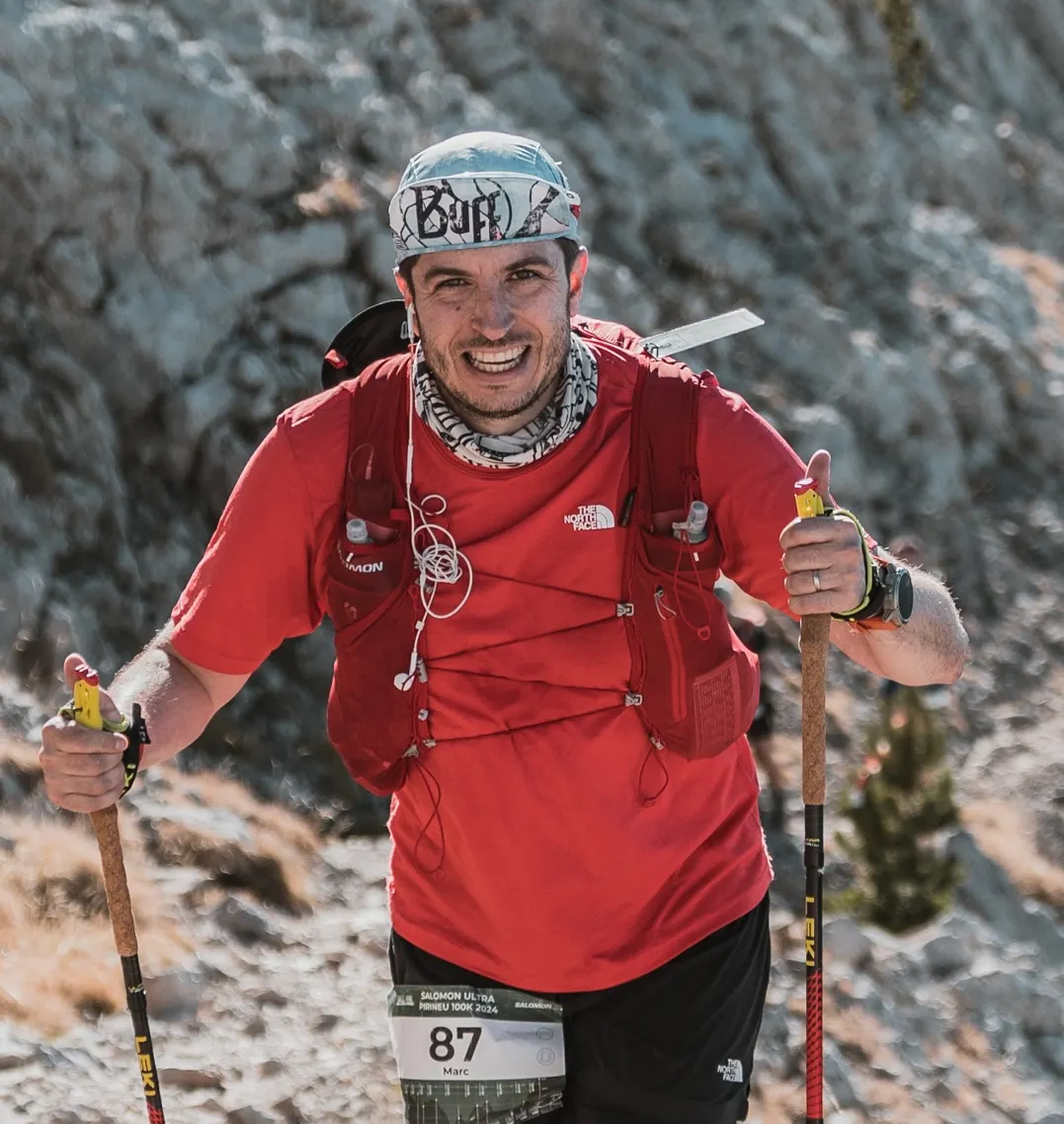 Marc smiling on a mountain trail with trekking poles and his red running kit